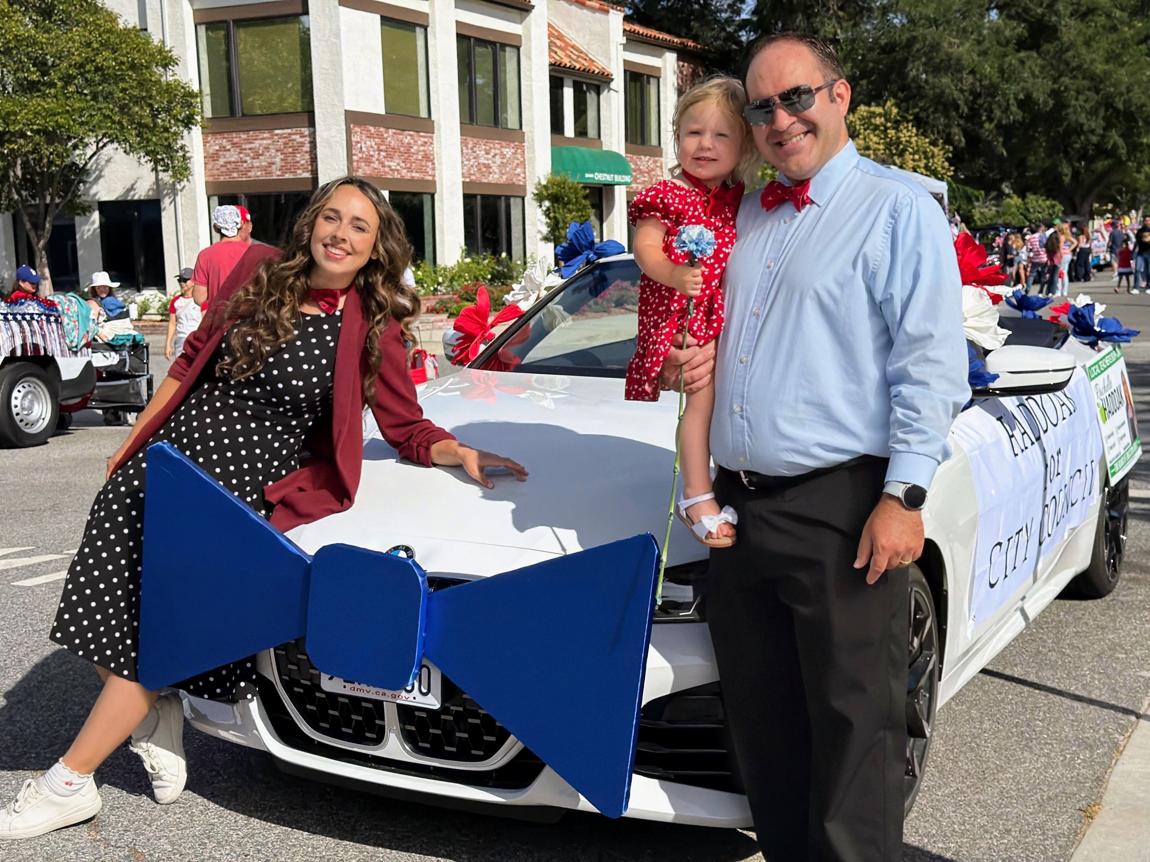 Brett Haddoak in a light blue shirt and red bow tie holding his daughter, standing with his wife Rachelle beside a white convertible decorated with a giant blue bow tie and a Haddoak for City Council sign.