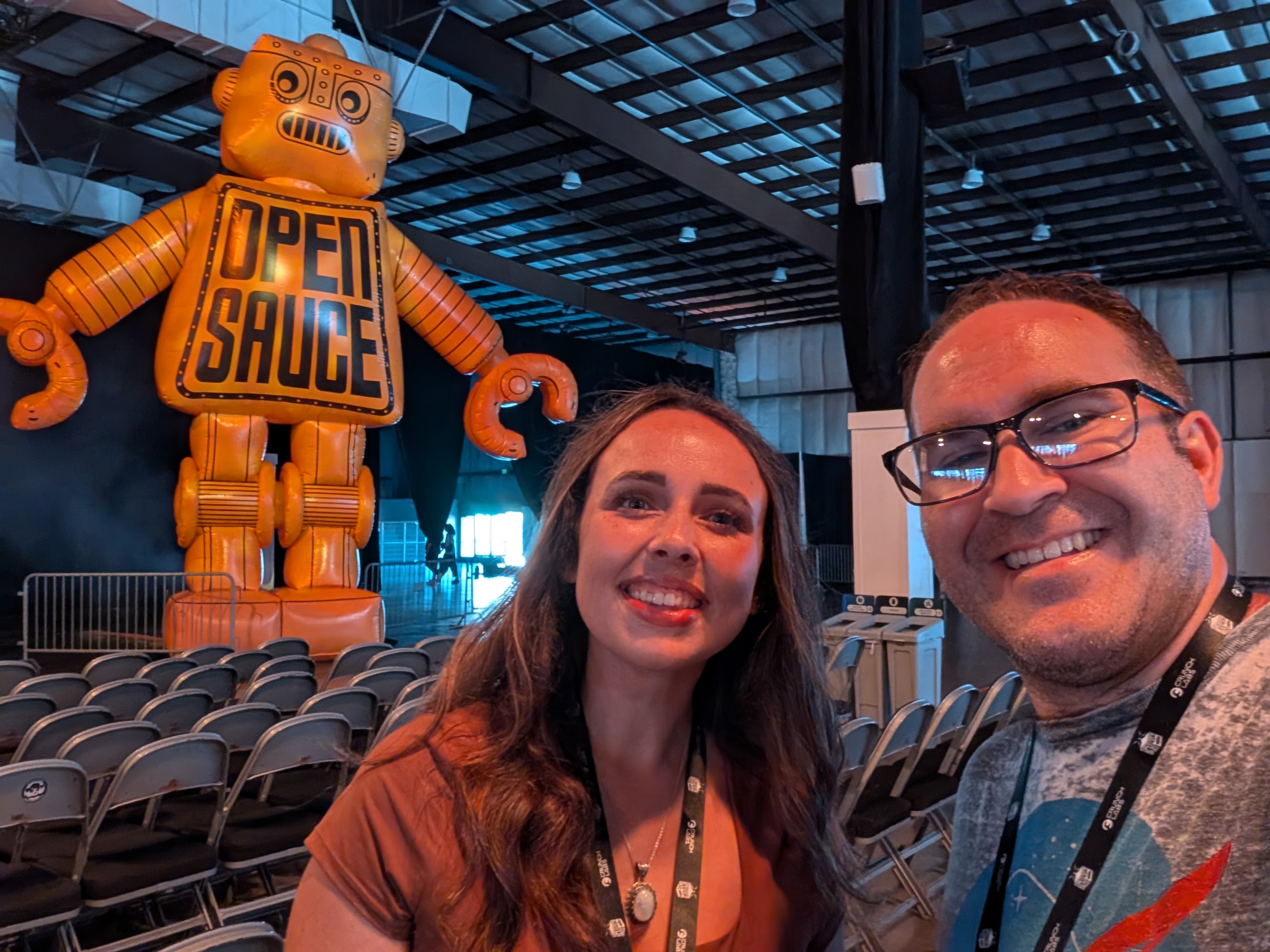 Selfie of Brett Haddoak and his wife Rachelle, both wearing event lanyards, in front of a giant inflatable orange Open Sauce robot.