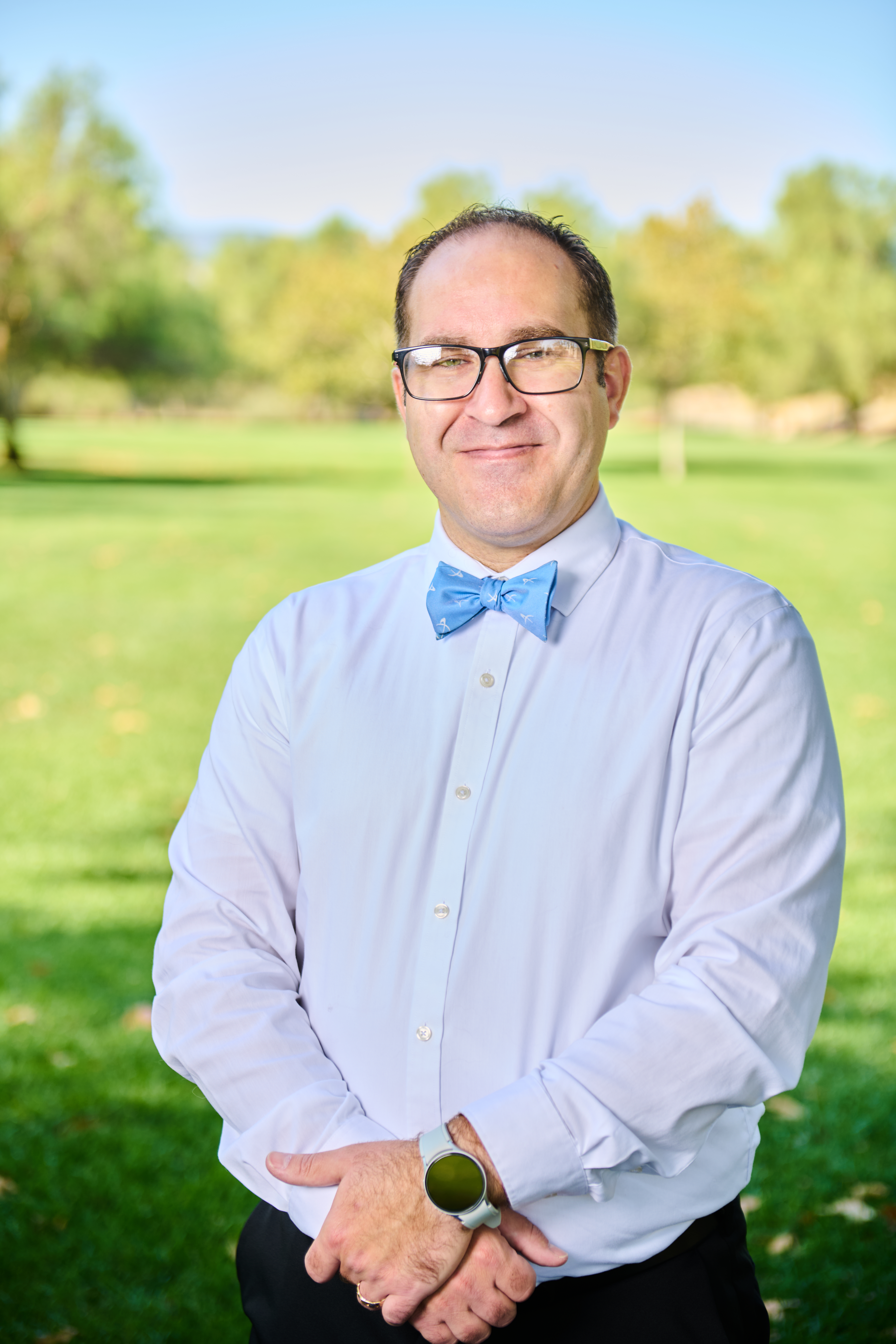 Brett Haddoak in a light blue dress shirt and blue bow tie, hands clasped in front, standing before a green park lawn.