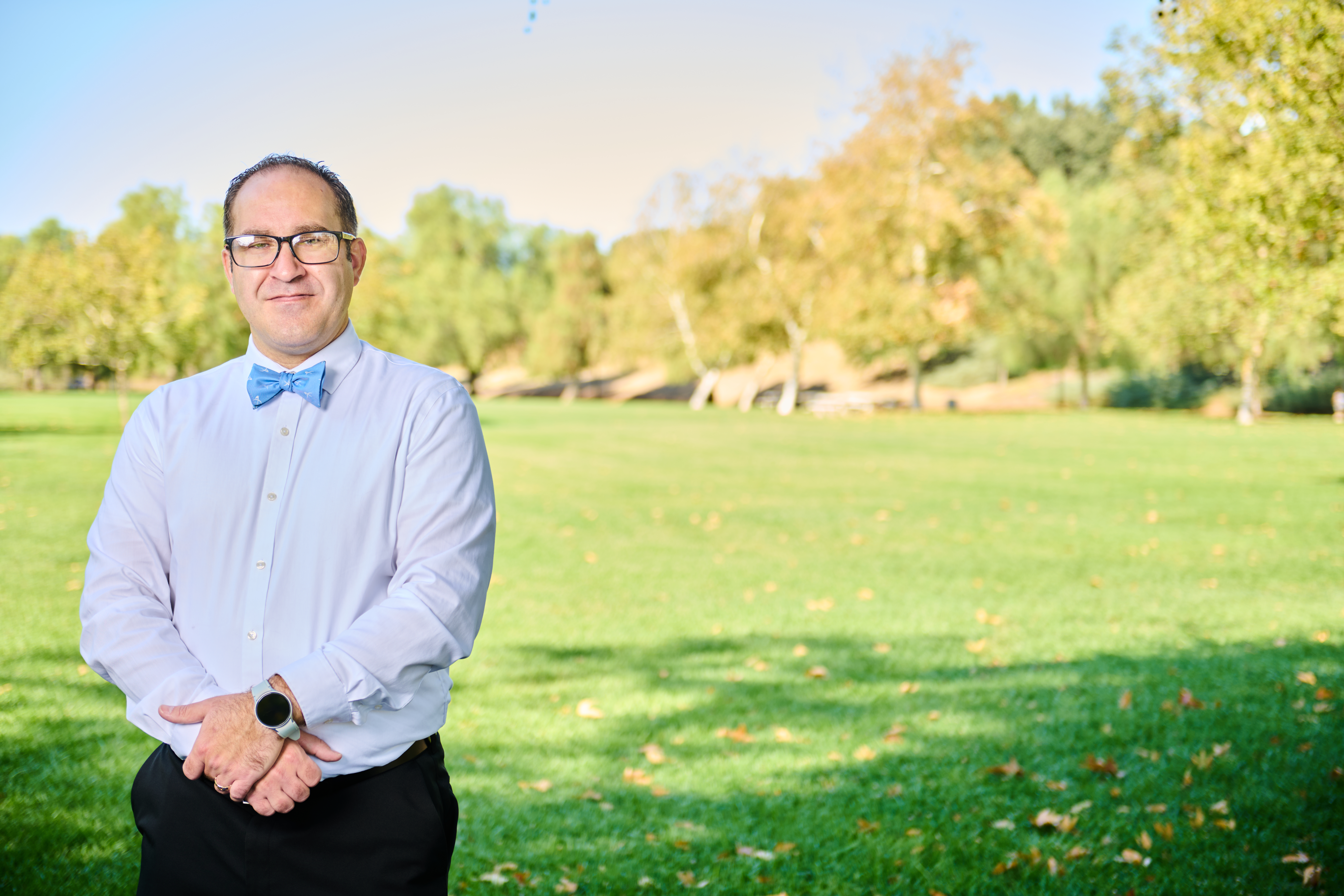 Brett Haddoak in a light blue dress shirt and blue bow tie, standing to the left of frame in a park with autumn foliage visible in the trees behind him.