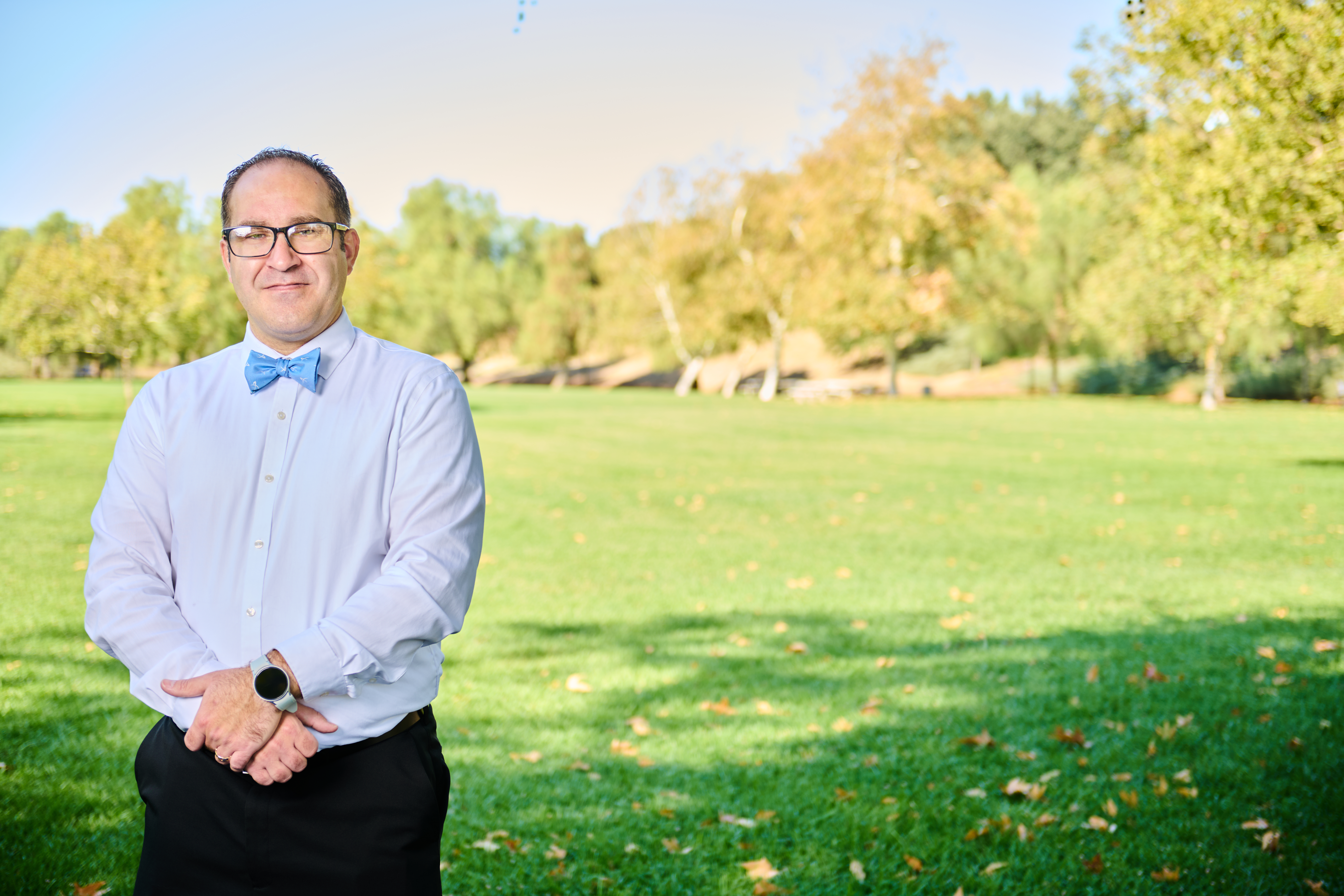Brett Haddoak in a light blue dress shirt and blue bow tie, standing to the left of frame before a park backdrop with warm autumn foliage.