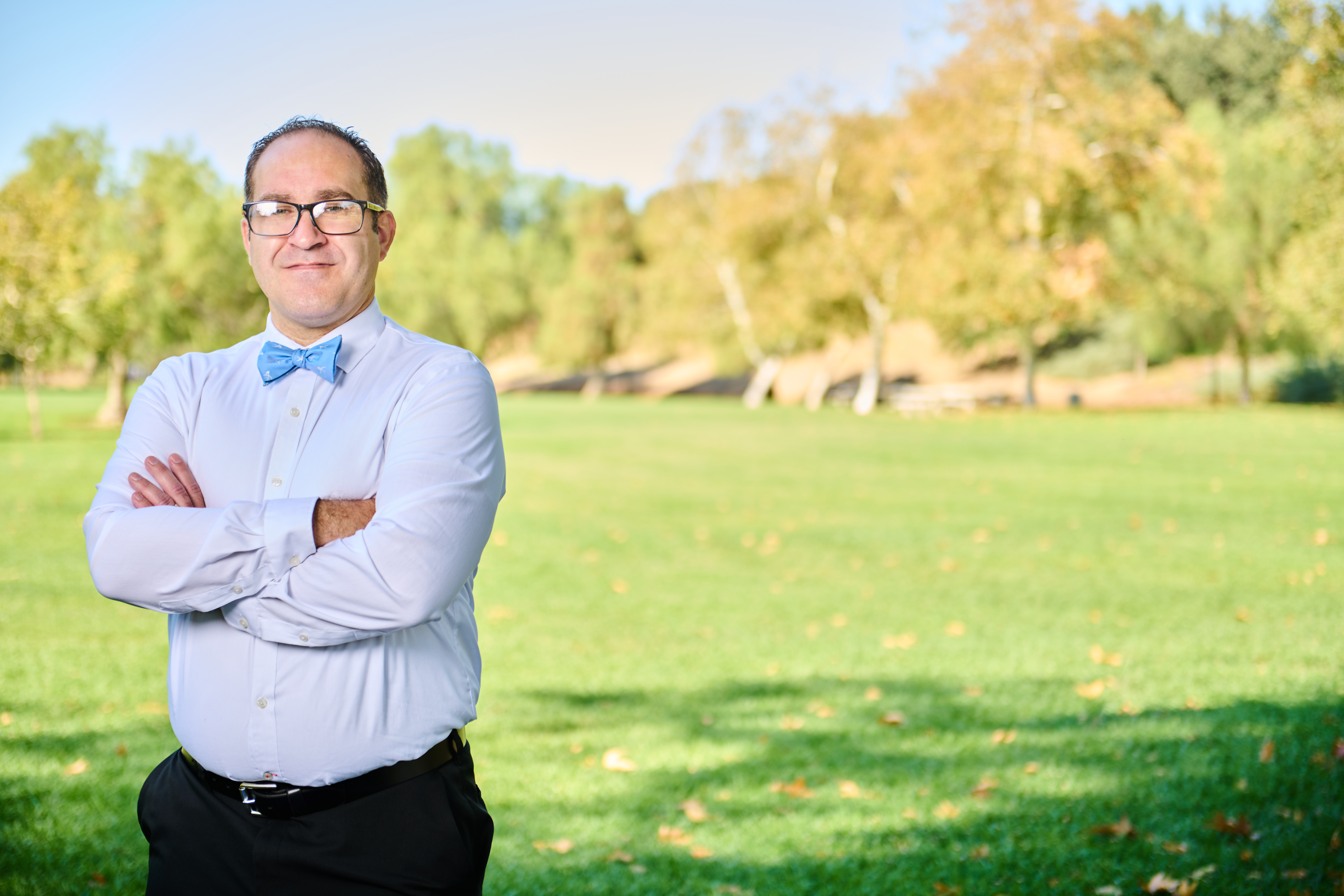 Brett Haddoak in a light blue dress shirt and blue bow tie, arms crossed, standing to the left of frame before a park with golden autumn trees.