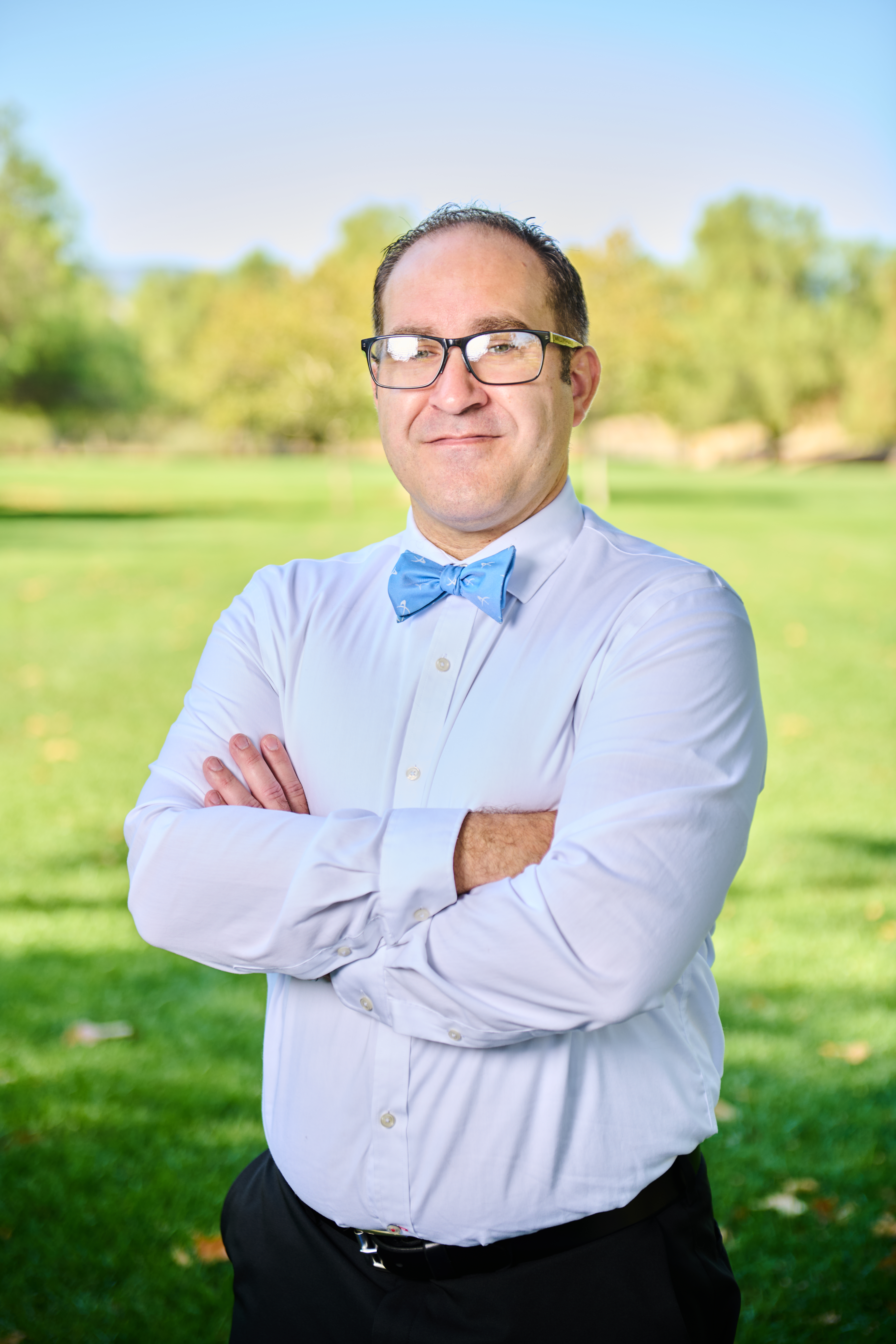 Brett Haddoak in a light blue dress shirt and blue bow tie, arms crossed, in a tighter crop against a green park background.