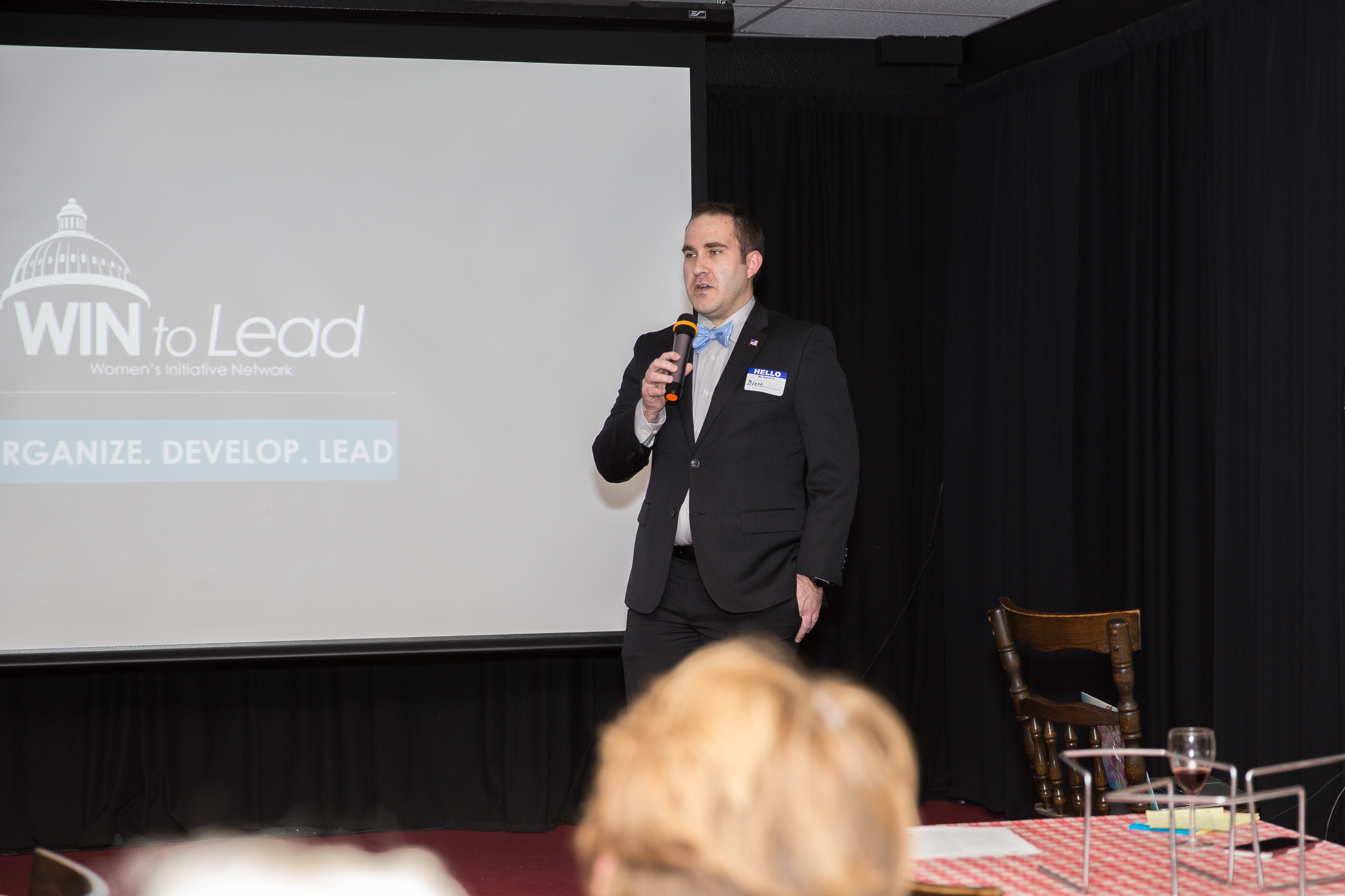 Brett Haddoak speaking into a handheld microphone beside a projected WIN to Lead slide.
